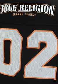 Czarny materiał z napisem "TRUE RELIGION BRAND JEANS" nad dużą białą siateczkową liczbą "02" obramowaną pomarańczowym ściegiem.