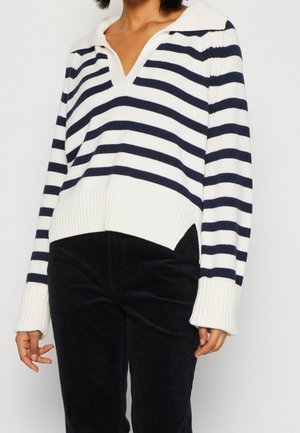 Femme portant un pull en tricot blanc et bleu marine à rayures avec un col en V et un pantalon en velours côtelé noir, se tenant devant un fond uni.