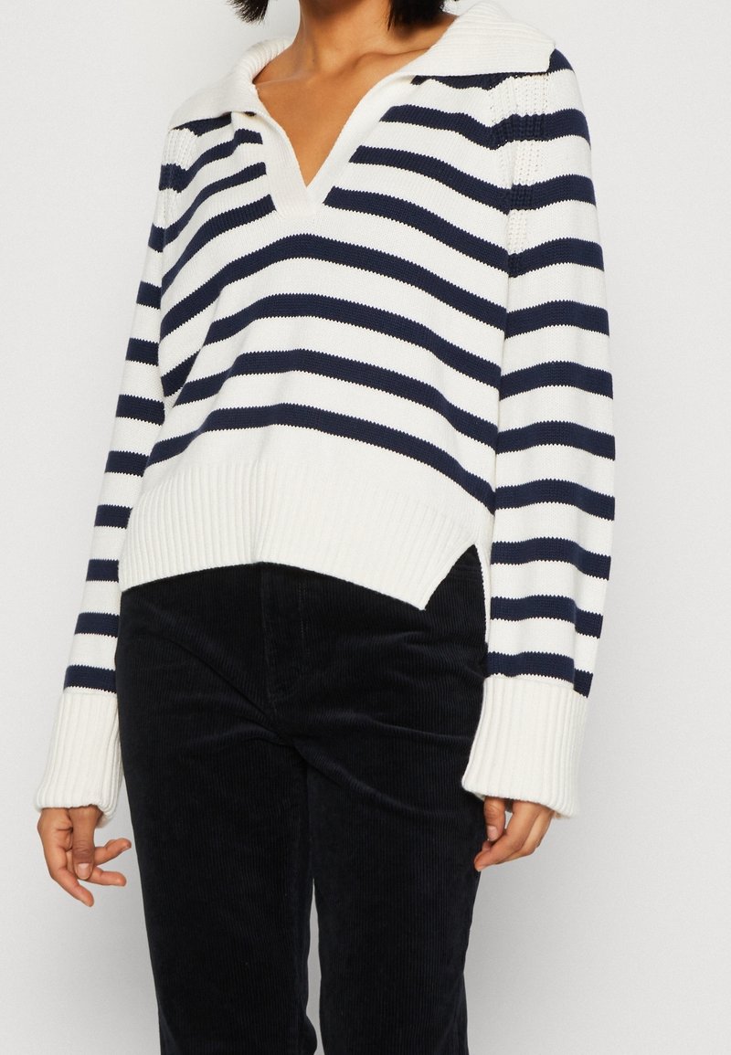 Femme portant un pull en tricot blanc et bleu marine à rayures avec un col en V et un pantalon en velours côtelé noir, se tenant devant un fond uni.