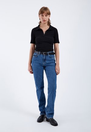 Młoda osoba stojąca, ubrana w czarną koszulkę z krótkim rękawem, niebieskie dżinsy z czarnym paskiem oraz czarne buty, na jednolitym jasnym tle.