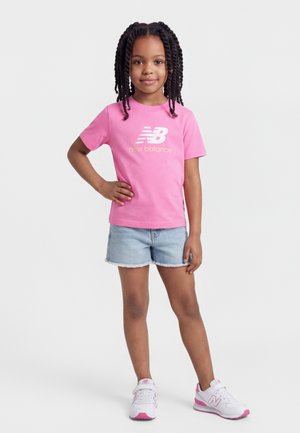 Mädchen mit geflochtenem Haar steht selbstbewusst da, trägt ein pinkes New Balance T-Shirt, Jeansshorts und weiße-pinke Sneakers vor weißem Hintergrund.