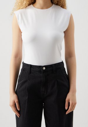 Vrouw draagt een mouwloze witte top met gewatteerde schouders en een zwarte broek met hoge taille, staand tegen een effen achtergrond.