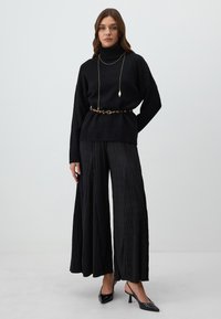 Un pull à col roulé noir associé à un pantalon large plissé noir et des escarpins noirs à bout pointu. Accentué par une ceinture en imprimé léopard et des bijoux en or.
