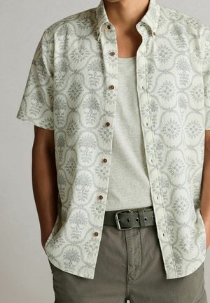 Homme portant une chemise à manches courtes boutonnée avec un motif floral clair sur un débardeur gris clair, associée à un pantalon gris et une ceinture foncée.