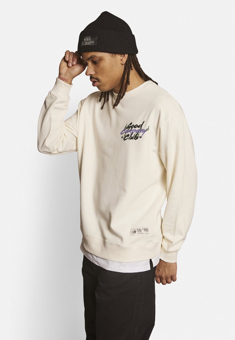 Sudadera de color crema con diseño bordado "Good Company" en púrpura y negro. Combínala con un gorro de punto negro. Parche sutil con el logotipo en el dobladillo.