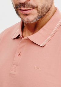 Koszulka polo w jasnoróżowym kolorze, wykonana z teksturowanej tkaniny, z guzikowym zapięciem i kontrastującymi detalami kołnierza. Logo wyszyte na piersi.