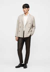 Νεαρός άνδρας στέκεται με τα χέρια στις τσέπες, φορώντας μπεζ σακάκι διπλότσεπο, λευκό πουκάμισο, σκούρο παντελόνι με σχέδιο και μαύρα επίσημα παπούτσια.