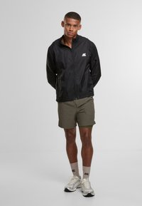 Giovane uomo in giacca zip nera, pantaloni corti verdi, calze beige e scarpe da ginnastica argentate in piedi contro uno sfondo grigio neutro.