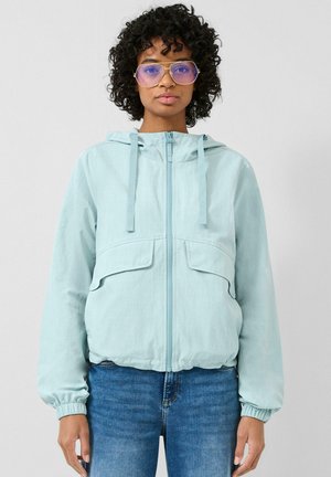 Kobieta z kręconymi włosami, nosząca duże okulary z fioletowym przyciemnieniem, jasnoniebieską bluzę z kapturem na zamek i niebieskie dżinsy, stojąca na jednolitym tle.