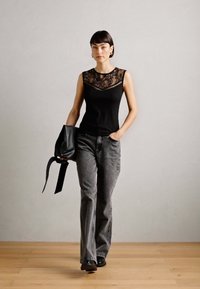 Melna bezpiedurkņu blūze ar mežģīņu detaļām ap kakla vietu, apvienota ar pelēkiem platām biksēm un melnajām kurpēm. Modelis tur melnu rokassomu.