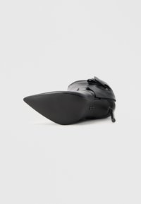 Escarpin noir à talon aigu avec un bout pointu et une tige en cuir brillant. Il présente un accent de nœud décoratif et un talon stiletto élancé.