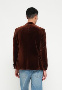 Man in een bruine fluwelen blazer en lichtblauwe jeans, met zijn rug naar de camera gekeerd tegen een effen witte achtergrond.