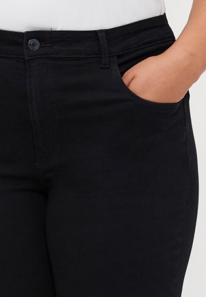 Zbliżenie osoby noszącej czarne spodnie z wysokim stanem, z ręką spoczywającą w przedniej prawej kieszeni.