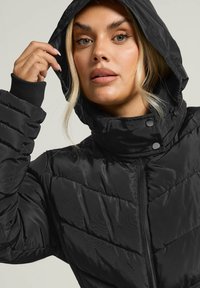 Schwarze Daunenjacke mit Reißverschluss, hohem Kragen und Kapuze. Texturiertes, gesteppte Design mit elastischen Bündchen und Druckknöpfen am Hals.