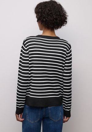 Person mit kurzen lockigen Haaren, der einen schwarz-weiß gestreiften Pullover mit langen Ärmeln und blaue Jeans trägt und einer hellgrauen Wand gegenübersteht.