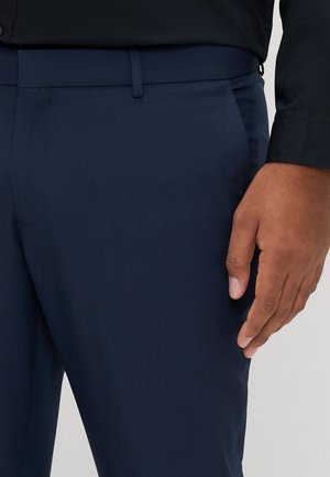 Pantalon bleu marine taillé dans un tissu lisse avec un devant plat, présentant une coupe ajustée et des poches latérales discrètes.