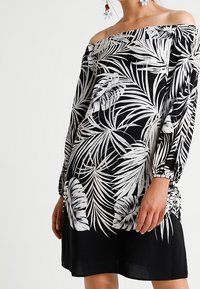 Olkaimattu musta mekko valkoisella trooppisella lehtikuvioinnilla, pitkät hihat ja rypytetty sivuosa, henkilö pukeutuneena kukkakoruihin.