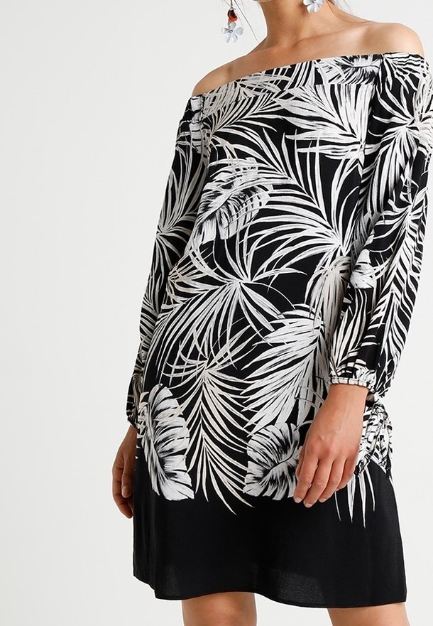 Olkaimattu musta mekko valkoisella trooppisella lehtikuvioinnilla, pitkät hihat ja rypytetty sivuosa, henkilö pukeutuneena kukkakoruihin.