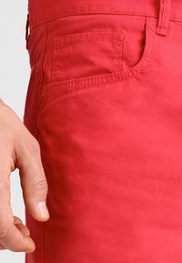 Red cotton shorts feature a side pocket with stitching details, highlighting the texture and color of the fabric. Hand positioned on the fabric.