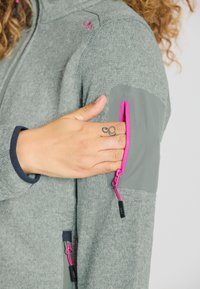 Fleece takki vaaleanharmaana, jossa on tummanharmaat kontrastipaneelit. Varustettu vaaleanpunaisilla vetoketjuviimeistelyillä ja vetoketjullisella taskulla käsivarressa.
