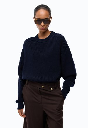Donna che indossa grandi occhiali da sole marroni, un maglione di lana blu scuro e pantaloni marroni scuri a vita alta con bottoni dorati.