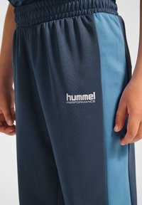 Marineblaue Sporthose mit einem türkisfarbenen Seitenstreifen; verfügt über einen elastischen Bund und das weiße Hummel Performance-Logo am Oberschenkel.