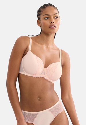 Woman with braided hair wearing a light peach bra and matching lace-trimmed panties, looking to the side against a plain background.