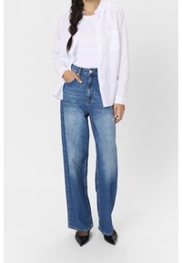 Jeans denim biru dengan pinggang tinggi dan potongan kaki lurus, menampilkan desain lima saku klasik dan sedikit pudar. Dipadukan dengan kemeja putih.