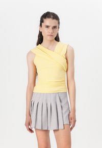 Top sans manches jaune avec un col tordu ; associé à une jupe plissée gris clair. Tissu lisse avec un design ajusté, peu de détails.