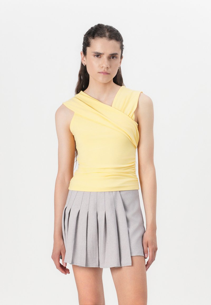 Top sans manches jaune avec un col tordu ; associé à une jupe plissée gris clair. Tissu lisse avec un design ajusté, peu de détails.