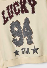 Krémová mikina s nápisom "LUCKY" v bordovej farbe, "94" v tmavosivej farbe a "STAR" so čiernymi hviezdičkami ako akcentovými detailmi. Mäkká, textúrovaná látka.