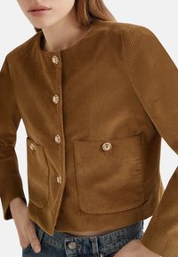 Giacca in corduroy marrone con scollatura rotonda, chiusura con bottoni frontali, due grandi tasche e trama a coste.