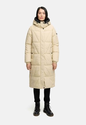 Beige gewatteerde lange jas met een hoge kraag en afneembare capuchon, met zwarte accenten en een gequilt ontwerp; gecombineerd met zwarte broek en laarzen.