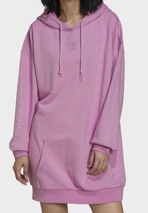 Femme portant une robe sweat à capuche oversize violet clair avec poche avant, cordons et logo en strass sur la poitrine et la manche.
