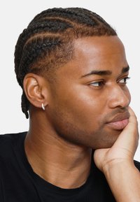 Cheveux noirs tressés avec une boucle d'oreille en argent. Peau lisse, expression neutre, portant un t-shirt noir, main reposant sur le menton.