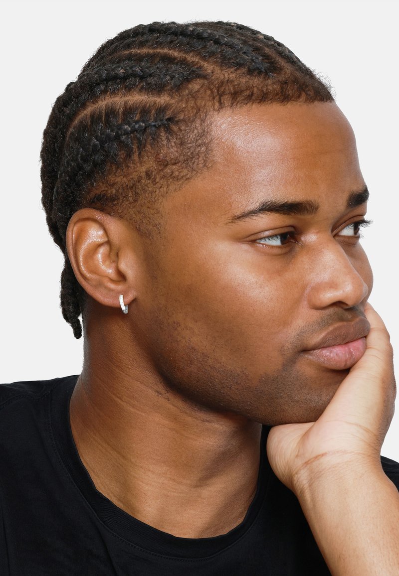 Cheveux noirs tressés avec une boucle d'oreille en argent. Peau lisse, expression neutre, portant un t-shirt noir, main reposant sur le menton.