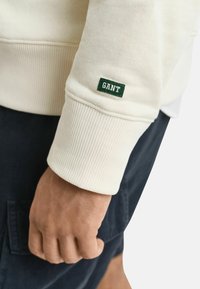 Crème-kleurig sweatshirt met geribbelde manchetten; heeft een groene GANT-logo label op de mouw gestikt; gecombineerd met donkere broek.