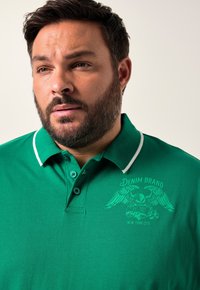 Grön polotröja med vita krageaccent, prydd med en broderad logotyp av en örn och text. Tillverkad av ett mjukt, bomullsliknande material.