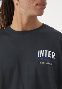 Dunkelgraues Baumwoll-T-Shirt mit kurzen Ärmeln. Mit einem aufgedruckten Logo in Weiß, Blau und goldener Schrift: "INTER Nerazzurri IM INTER, IM MILANO."