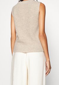 Femme portant un gilet beige sans manches en tricot et un pantalon blanc ample, vue de dos sur un fond uni.