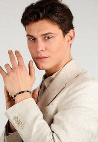 Svart armbandsur på modellens handled som hålls av höger hand, med cirkulära accenter. Modellen bär en ljusbeige kavaj med knappar. Slät hudtextur.