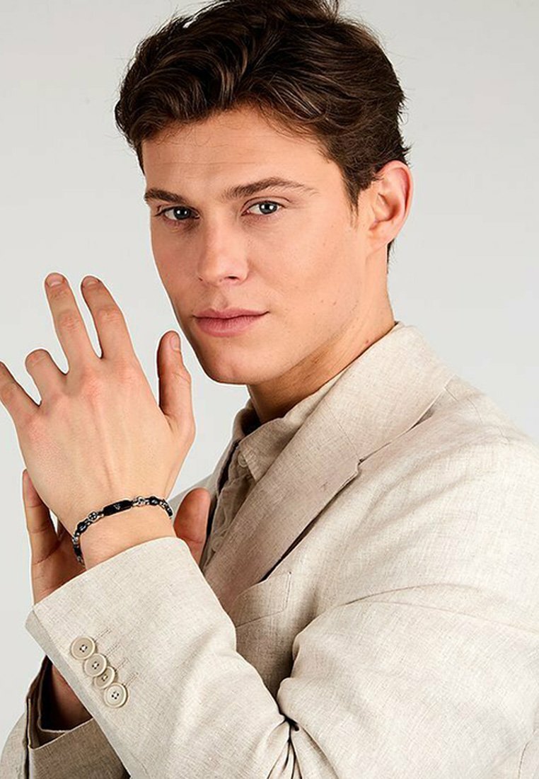 Svart armbandsur på modellens handled som hålls av höger hand, med cirkulära accenter. Modellen bär en ljusbeige kavaj med knappar. Slät hudtextur.