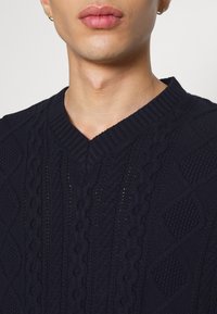 Pull en tricot bleu marine avec un col en V, présentant des motifs en diamants texturés et en câble. Vue rapprochée mettant en valeur les détails des points et de l'encolure.