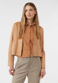 Giovane donna con lunghi capelli biondi, che indossa una giacca in camoscio beige con zip, una camicia marrone con bottoni e pantaloni beige a quadri, in piedi davanti a uno sfondo bianco.