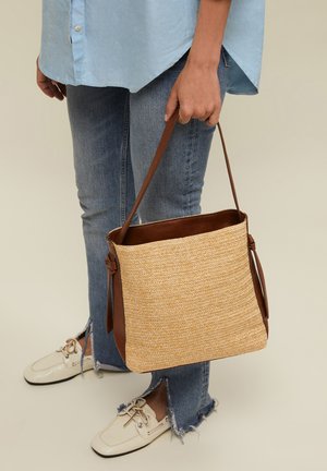 Inimene, kes kannab helesinist särki ja kulunud äärega teksaseid, hoiab käes tan värvi kootud käekotti pruunide nahkrihmadega ja kreemjad paeltega loafers'eid.