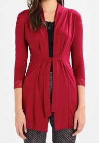 Červený kardigan s šálovým límcem a tříčtvrtečními rukávy, s krajkovými detaily na spodním lemu a manžetách, uvázaný v pase.
