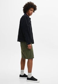 Jeune homme aux cheveux bouclés portant une veste noire, un short vert olive, des chaussettes blanches et des baskets noires, debout de profil sur un fond blanc.
