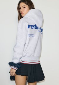 Grijze capuchontrui met blauwe bedrukte tekst en gestreepte accenten aan de zoom. Zwarte geplooide rok maakt de outfit compleet.