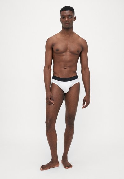 Hvide menns briefs med en sort elastisk talje, der fremhæver en glat stofstruktur og en tætsiddende design på en model med atletisk fysik.