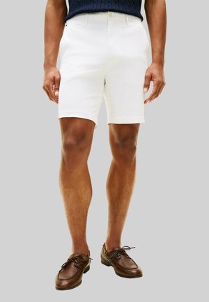 Uomo che indossa pantaloncini bianchi su misura e scarpe stringate in pelle marrone davanti a uno sfondo grigio uniforme.